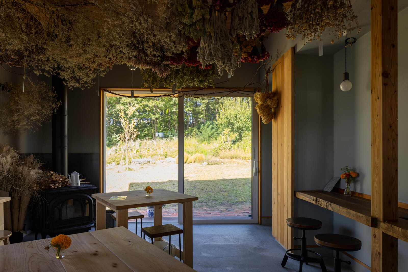 庭との一体感、動線の良さを実現する大開口を設けた店舗スペース。掃除のしやすい土間仕上げの床には、ヨツールの薪ストーブも設置。木とモルタル、珪藻土で仕上げたシンプルな箱のような店内を、奥さんが育てた色鮮やかな花々と多肉植物が彩る