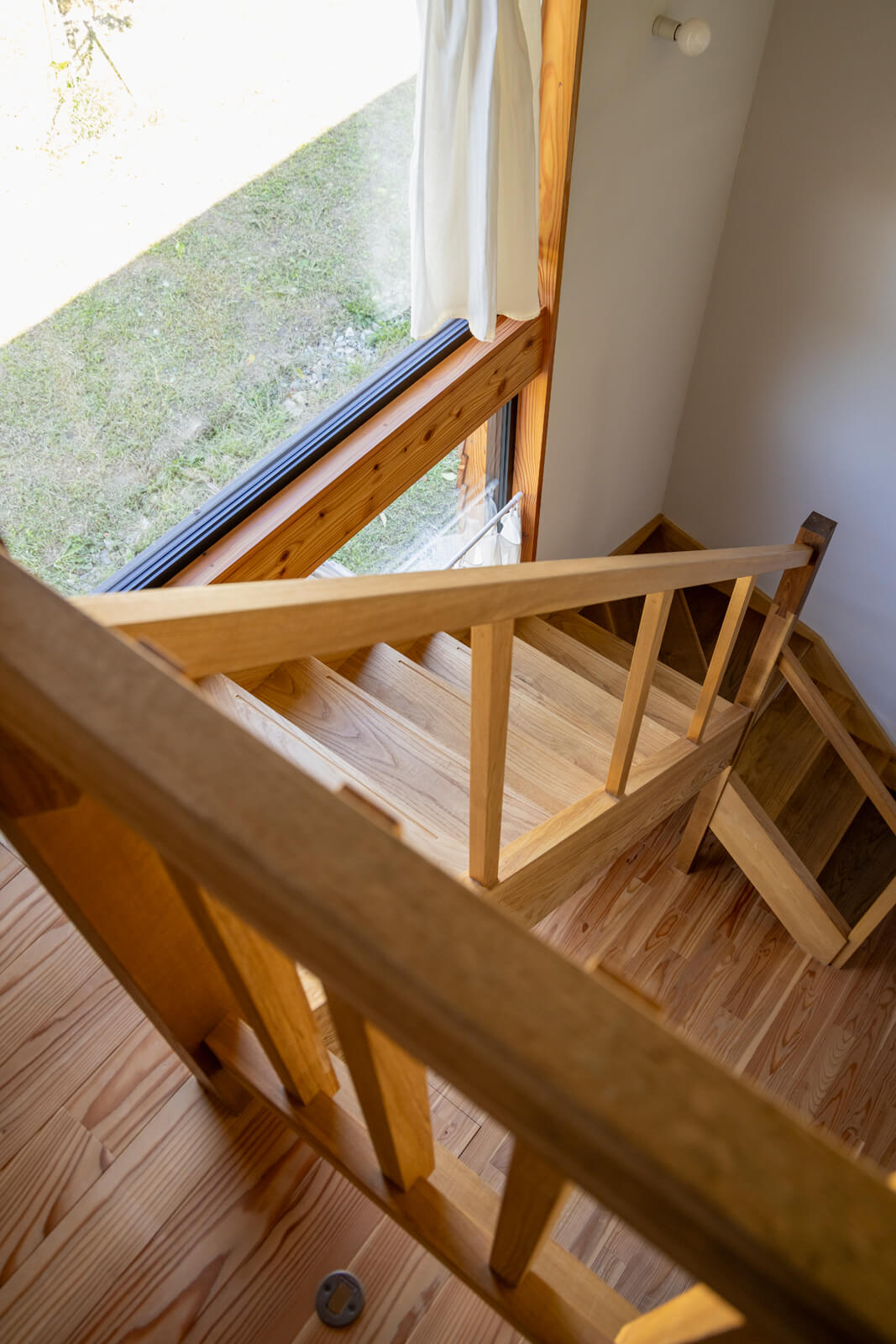 居住スペースの階段は、吹き抜けの3連窓からの自然光がきれいに射すようにスケルトン仕上げに。武部建設でストックしていたナラとタモ、キハダ、エンジュの無垢材を使用し、親柱の婆娑羅（ばさら）継ぎという伝統手法で材をつなぎ合わせて意匠とした